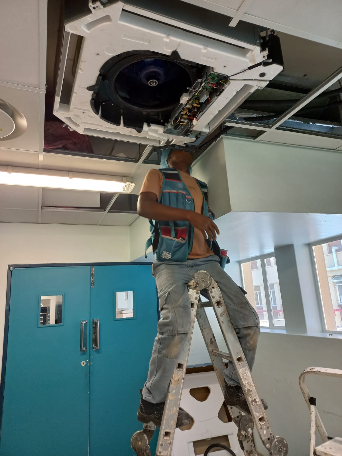 Technician on a ladder servicing a ceiling cassette air conditioning unit in a commercial building